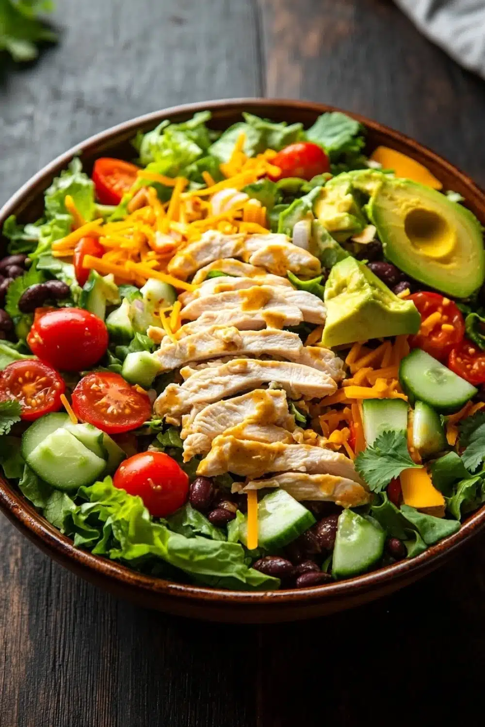 Chopped chicken salad with creamy chipotle dressing - the image is of a large bowl of salad on a wooden table. the salad is made up of various ingredients such as lettuce, tomatoes, black beans, sliced avocado, shredded cheese, and cilantro. the ingredients are arranged in a colorful and appetizing manner. the bowl is brown and appears to be made of ceramic or ceramic. the background is blurred, but it seems to be a kitchen countertop with a few sprigs of parsley scattered around.