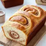 Cinnamon bread - the image shows a freshly baked cinnamon roll on a wooden cutting board. the roll is golden brown in color and has a swirl pattern on top. it appears to be freshly baked and is sitting on a white marble countertop. there are two small bowls of dipping sauce on the cutting board next to the roll. the background is blurred, but it seems to be a kitchen countertop with more rolls of bread in the background.