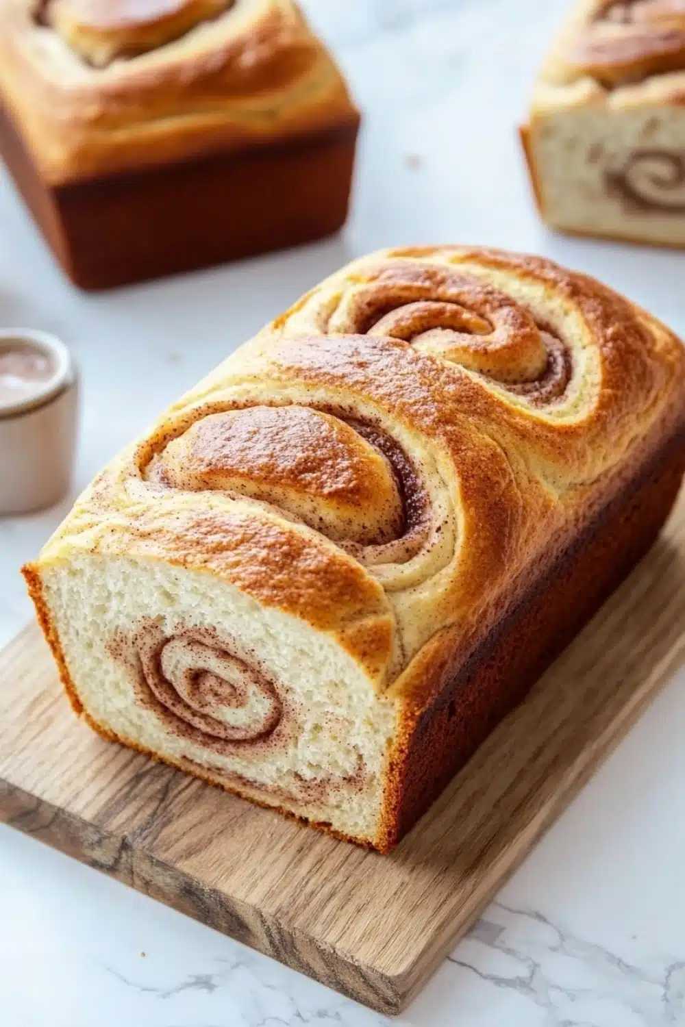 Cinnamon bread - the image shows a freshly baked cinnamon roll on a wooden cutting board. the roll is golden brown in color and has a swirl pattern on top. it appears to be freshly baked and is sitting on a white marble countertop. there are two small bowls of dipping sauce on the cutting board next to the roll. the background is blurred, but it seems to be a kitchen countertop with more rolls of bread in the background.