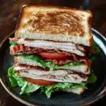 Club sandwich - the image shows a sandwich on a black plate. the sandwich is made with two slices of toasted bread, filled with sliced turkey, bacon, and lettuce. the turkey is cut into thin slices and the bacon is cooked to a golden brown color. the lettuce is a mix of green and red, and the tomato is a bright red. the plate is sitting on a wooden table with a pair of chopsticks resting on the side.