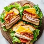 Club sandwich on french bread - the image shows a wooden cutting board with three sandwiches on it. the sandwiches are made with two slices of toasted bread, filled with sliced ham, cheese, and lettuce. the ham is cut into thin slices and is arranged in a circular pattern on top of the cheese. the cheese is melted and bubbly, and the lettuce is a vibrant green color. the cutting board is placed on a white marble countertop.