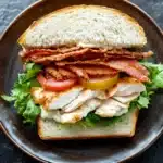 Club sandwich with chicken - the image is of a sandwich on a brown plate. the sandwich is made with a toasted white bread and is filled with a variety of ingredients. on top of the bread, there are strips of bacon, sliced tomatoes, and a slice of yellow bell pepper. the bacon is crispy and golden brown, and the tomatoes are bright red. the lettuce is a mix of green and yellow, and there is a layer of white sauce on top. the plate is sitting on a dark grey textured surface.