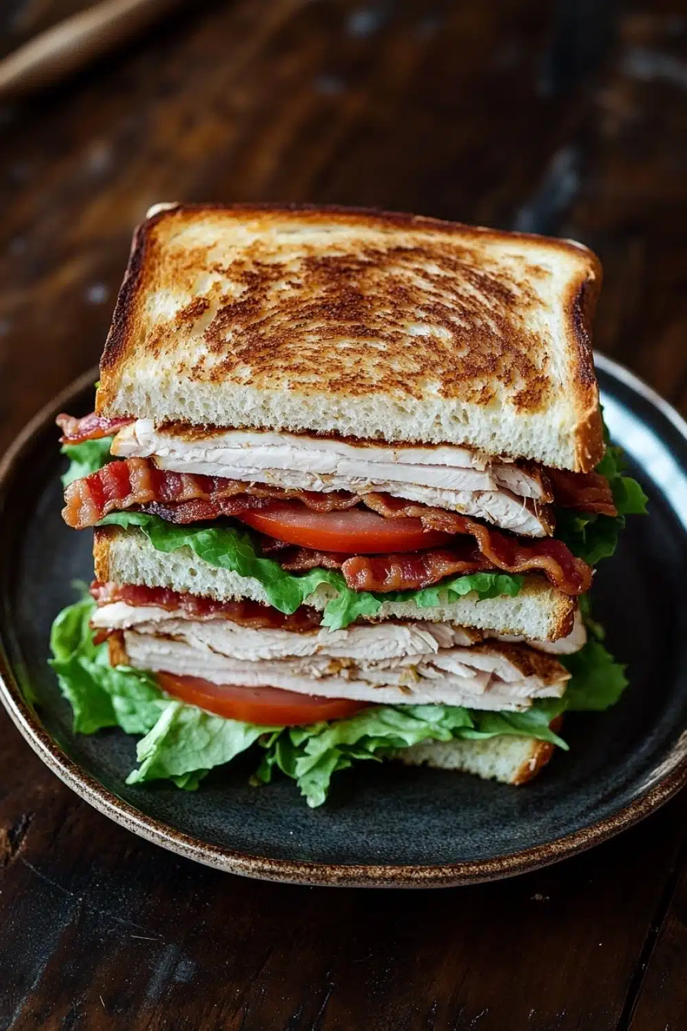 Club sandwich - the image shows a sandwich on a black plate. the sandwich is made with two slices of toasted bread, filled with sliced turkey, bacon, and lettuce. the turkey is cut into thin slices and the bacon is cooked to a golden brown color. the lettuce is a mix of green and red, and the tomato is a bright red. the plate is sitting on a wooden table with a pair of chopsticks resting on the side.