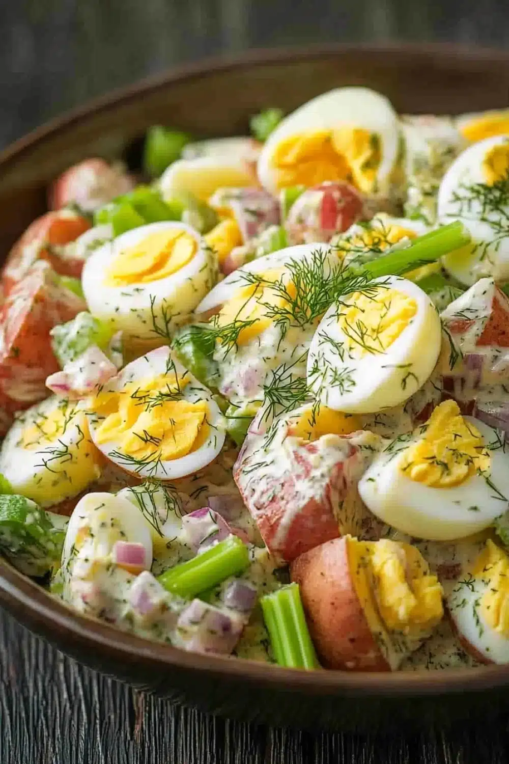 Creamy dill red potato salad - the image is a close-up of a bowl of potato salad. the salad is made up of diced potatoes, diced red onions, and hard-boiled eggs. the potatoes are arranged in a circular pattern, with some of them overlapping each other. the eggs are cut in half and are topped with a dollop of dill. the dish is garnished with a sprig of fresh dill, adding a pop of green color to the dish. the bowl is sitting on a wooden table, and the background is blurred, making the salad the focal point of the image.
