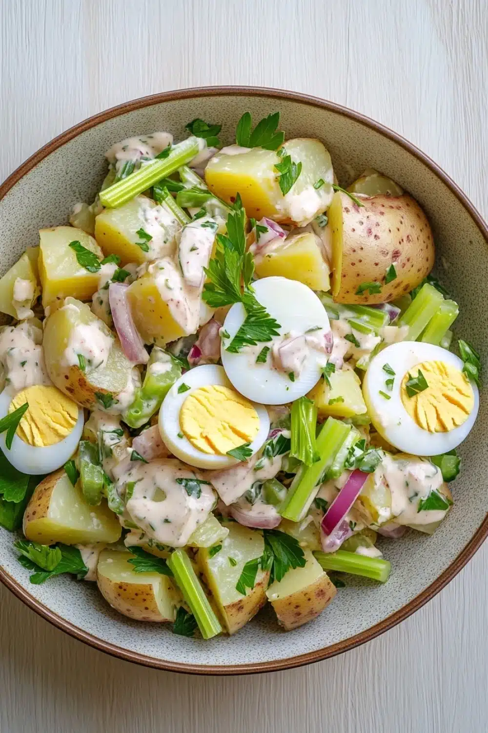 Creamy potato and egg salad - the image is a close-up of a bowl of potato salad. the bowl is made of light-colored ceramic and is placed on a white wooden table. the salad is made up of diced potatoes, diced celery, red onions, and hard-boiled eggs. the potatoes are arranged in a pile and are topped with a creamy dressing. the dressing is drizzled over the top of the potatoes and is garnished with fresh parsley. the overall appearance of the salad is colorful and appetizing.