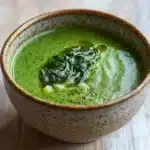 Creamy soup with spinach - the image is a close-up of a bowl of green soup on a wooden table. the soup appears to be a creamy green color and has a smooth texture. on top of the soup, there is a small amount of fresh spinach leaves, which are a vibrant green color. the bowl is made of ceramic and has speckled edges. the background is blurred, making the bowl the focal point of the image.