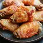 Crispy chicken wings without frying - the image shows a plate of grilled chicken wings. the wings are golden brown and appear to be seasoned with herbs and spices. they are arranged in a circular pattern on the plate, with some overlapping each other. the plate is dark brown and has a textured surface. the background is blurred, but it appears to be a wooden table or countertop.