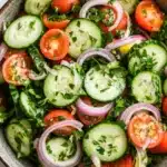 Cucumber tomato onion salad lemon - the image is a close-up of a bowl of salad. the salad is made up of sliced cucumbers, red onions, cherry tomatoes, and cilantro. the cucumbers are cut into thin slices and are arranged in a circular pattern. the tomatoes are bright red and appear to be ripe and juicy. the onions are sliced into thin strips and are scattered throughout the salad, with some of them overlapping each other. the cilantro is finely chopped and adds a pop of green color to the dish. the bowl is made of a light-colored ceramic or ceramic material and is sitting on a white marble countertop.