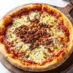 Deep dish pizza premade dough - the image is of a freshly baked pizza on a wooden cutting board. the pizza is round and has a golden brown crust. it is topped with melted cheese, red sauce, and chunks of ground meat. there are also some herbs scattered on top of the cheese. on the right side of the cutting board, there is a small jar of black peppercorns. the cutting board is placed on a white marble countertop.