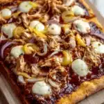 Detroit style bbq chicken pizza - the image is of a square pizza on a wooden cutting board. the pizza has a golden brown crust and is topped with shredded chicken, sliced onions, and mozzarella cheese. the chicken is cooked to a medium-rare and appears to be seasoned with herbs and spices. the cheese is melted and bubbly, and the sauce is a deep red color. the cutting board is placed on a white wooden table.
