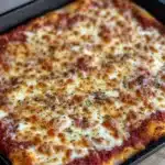 Detroit style pizza in cast iron - the image shows a rectangular baking tray with a freshly baked pizza in it. the pizza has a golden brown crust and is topped with melted cheese and red sauce. the cheese is melted and bubbly, and there are small black pepper flakes scattered throughout the top. the tray is sitting on a wooden cutting board. the background is blurred, but it appears to be a kitchen countertop.
