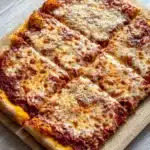 Detroit style pizza with frozen bread dough - the image shows a square pizza on a wooden cutting board. the pizza has a golden brown crust and is topped with melted cheese and tomato sauce. the cheese is golden brown and appears to be melted and bubbly. the tomato sauce is a deep red color and is spread evenly across the top of the pizza. the cutting board is placed on a white wooden table.