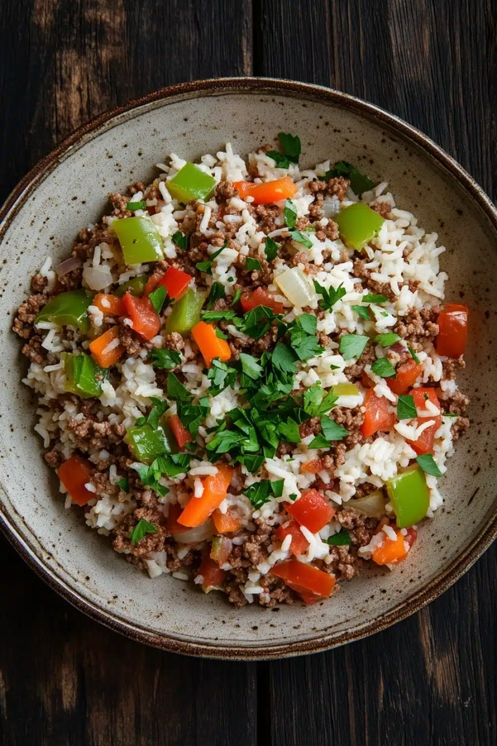Dinner with ground beef and rice - the image is a close-up of a bowl of food on a wooden table. the bowl is made of ceramic and has a light brown color. the food in the bowl appears to be a mixture of rice, ground beef, diced tomatoes, and green bell peppers. the rice is white and fluffy, and there are small pieces of minced meat scattered throughout the dish. the vegetables are bright red and appear to be seasoned with herbs and spices. the dish is garnished with a sprig of fresh parsley. the overall appearance of the dish is colorful and appetizing.