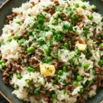 Garlic butter beef rice skillet - the image is a close-up of a dish in a black bowl. the dish appears to be a type of rice or risotto, with chunks of ground beef and peas mixed in. the rice is white and fluffy, and the peas are bright green. there are also small pieces of diced potatoes scattered throughout the dish, adding a pop of color and texture. the bowl is garnished with a sprig of fresh parsley. the background is a dark grey countertop, and there are a few sprigs of parsley scattered around the bowl.