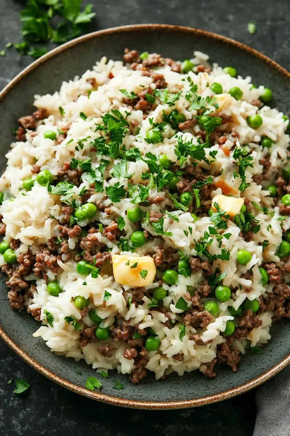 Garlic butter beef rice skillet - the image is a close-up of a dish in a black bowl. the dish appears to be a type of rice or risotto, with chunks of ground beef and peas mixed in. the rice is white and fluffy, and the peas are bright green. there are also small pieces of diced potatoes scattered throughout the dish, adding a pop of color and texture. the bowl is garnished with a sprig of fresh parsley. the background is a dark grey countertop, and there are a few sprigs of parsley scattered around the bowl.