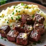 Garlic butter steak bites and mash - the image shows a plate of food with a serving of mashed potatoes. the plate is brown and has a speckled pattern on it. on top of the mashed potatoes, there are several pieces of cooked beef cubes arranged in a circular pattern. the beef cubes are dark brown and appear to be seasoned with herbs and spices. the mashed potatoes are creamy and fluffy, and there are a few sprigs of parsley scattered around the plate. the background is blurred, but it appears to be a wooden table with a basket of herbs on the side.