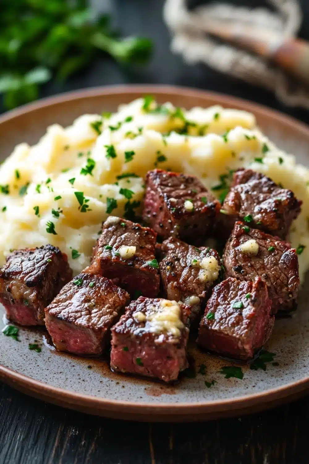 Garlic butter steak bites and mash - the image shows a plate of food with a serving of mashed potatoes. the plate is brown and has a speckled pattern on it. on top of the mashed potatoes, there are several pieces of cooked beef cubes arranged in a circular pattern. the beef cubes are dark brown and appear to be seasoned with herbs and spices. the mashed potatoes are creamy and fluffy, and there are a few sprigs of parsley scattered around the plate. the background is blurred, but it appears to be a wooden table with a basket of herbs on the side.