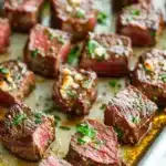 Garlic butter steak bites chuck roast - the image shows a baking tray with several pieces of cooked steak on it. the steak appears to be medium-rare and has a pinkish-red color. it is garnished with chopped herbs and a sprinkle of herbs on top. the tray is lined with parchment paper and there is a drizzle of sauce on the side. the background is blurred, but it seems to be a kitchen countertop.