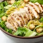Gluten free chicken caesar pasta salad - the image is a close-up of a bowl of pasta salad. the bowl is made of ceramic and has a brown rim. the salad is made up of green leafy vegetables, possibly lettuce, and is topped with sliced chicken and croutons. the chicken is cooked to a golden brown color and appears to be seasoned with herbs and spices. the pasta is fusilli, which is a type of spiral-shaped pasta, and the salad is drizzled with a creamy dressing. the background is blurred, but it seems to be a white tablecloth.