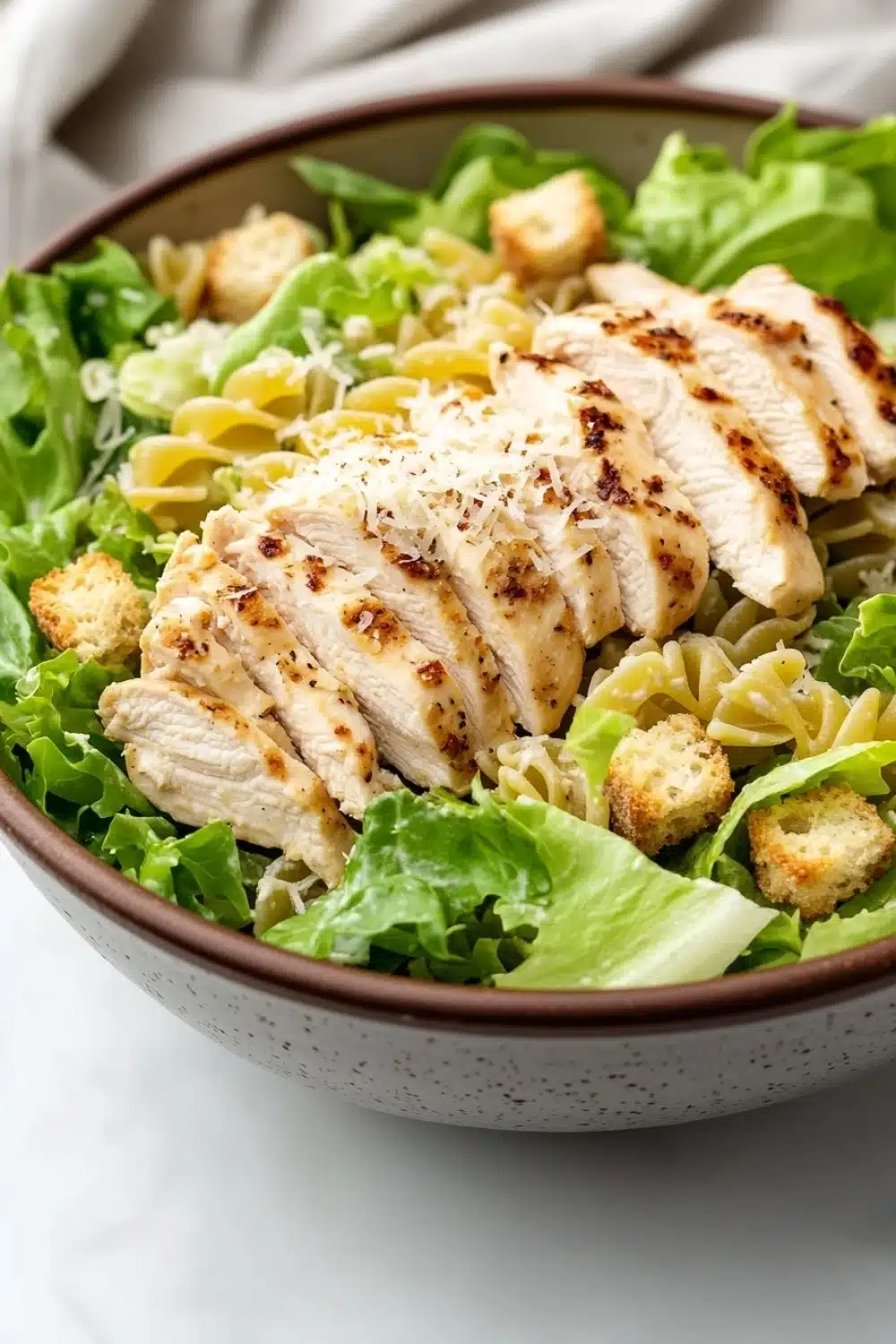 Gluten free chicken caesar pasta salad - the image is a close-up of a bowl of pasta salad. the bowl is made of ceramic and has a brown rim. the salad is made up of green leafy vegetables, possibly lettuce, and is topped with sliced chicken and croutons. the chicken is cooked to a golden brown color and appears to be seasoned with herbs and spices. the pasta is fusilli, which is a type of spiral-shaped pasta, and the salad is drizzled with a creamy dressing. the background is blurred, but it seems to be a white tablecloth.