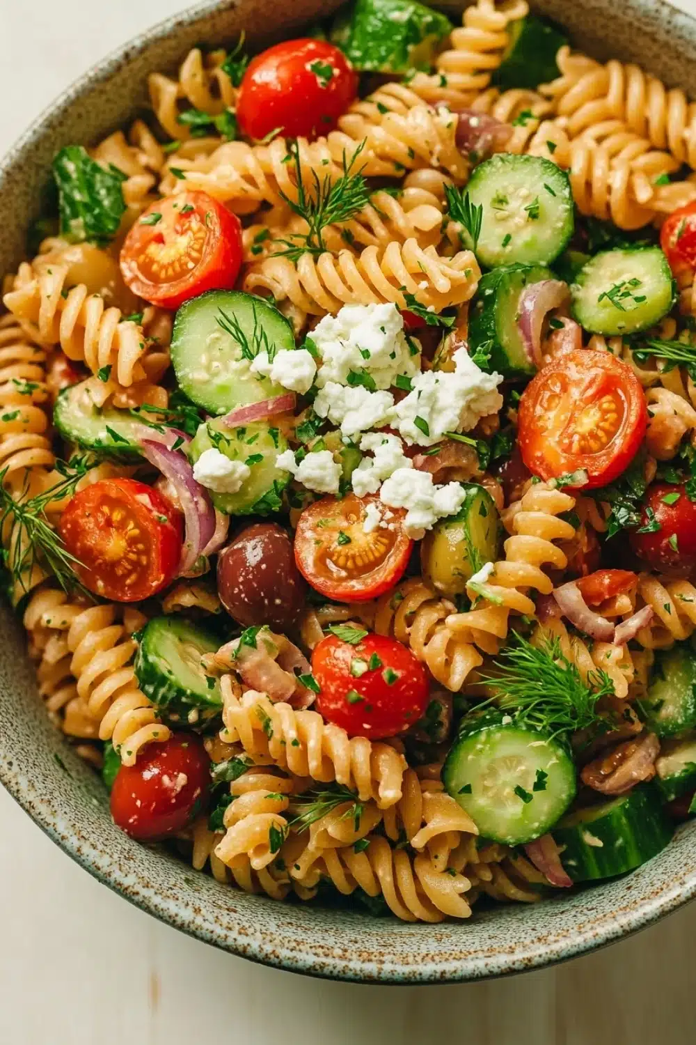 Greek pasta salad vegan - the image is a close-up of a bowl of pasta salad. the bowl is made of ceramic and has a blue and white speckled pattern. the pasta is fusilli, which is a type of spiral-shaped pasta, and is mixed with various vegetables such as sliced tomatoes, cucumbers, red onions, and olives. the vegetables are garnished with fresh dill and crumbled feta cheese. the salad appears to be freshly prepared and ready to eat.