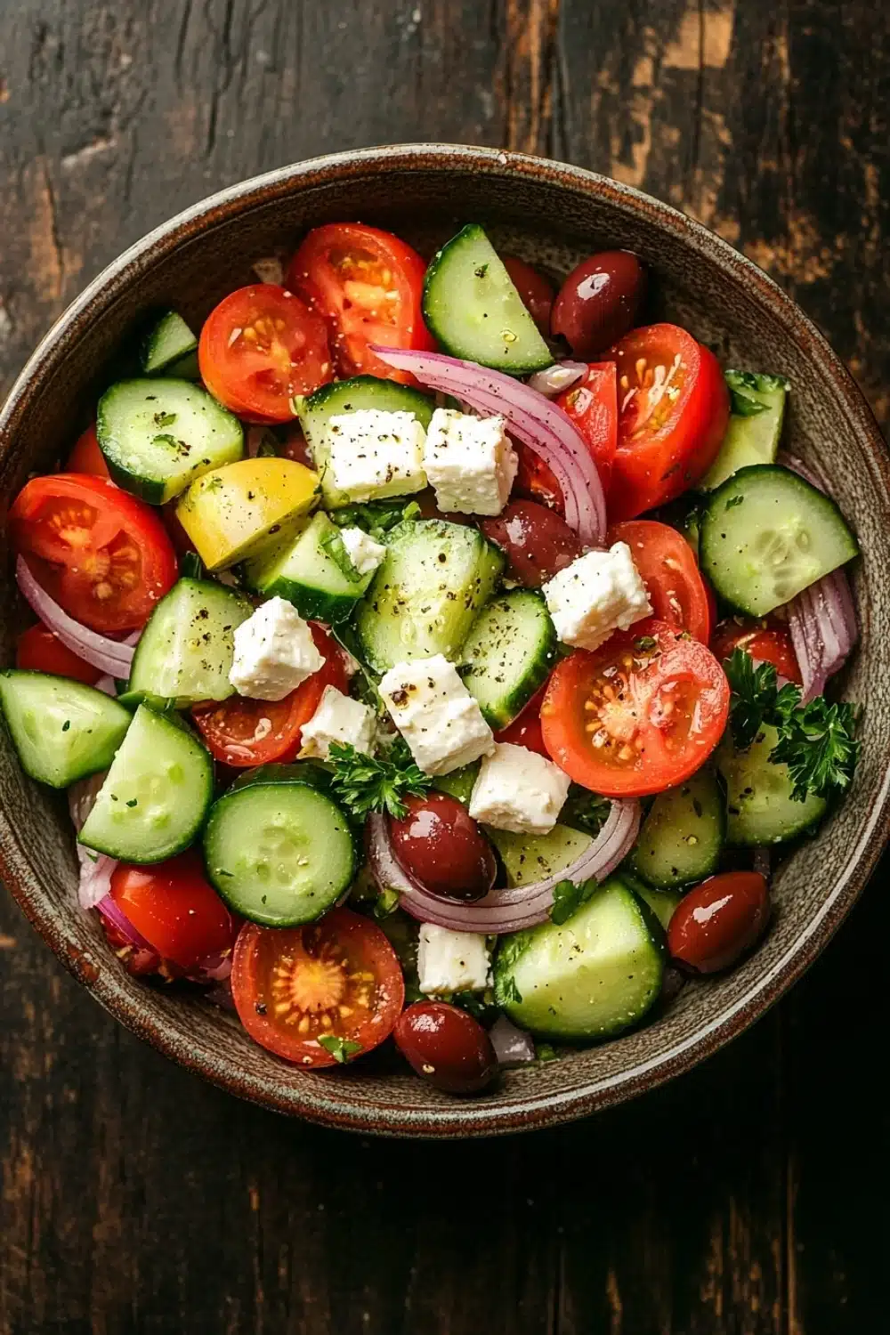 Greek salad - the image is a close-up of a bowl of salad on a wooden table. the salad is made up of sliced cucumbers, cherry tomatoes, red onions, and feta cheese. the cucumbers are cut into thin slices and are arranged in a circular pattern. the cherry tomatoes are bright red and appear to be ripe and juicy. the red onions are sliced into thin strips and are scattered throughout the salad. the feta is crumbled and adds a creamy texture to the dish. there are also some sprigs of parsley on top of the salad, adding a pop of green color. the bowl is made of dark wood and has a rustic, weathered appearance.
