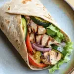 Grilled chicken and veggie wrap - the image shows a wrap on a blue plate. the wrap is made of tortilla and is filled with grilled chicken, zucchini, red onion, and cherry tomatoes. the chicken is cooked to a golden brown color and appears to be seasoned with herbs and spices. the vegetables are arranged in a circular pattern around the wrap, with some overlapping each other. the plate is sitting on a wooden table.
