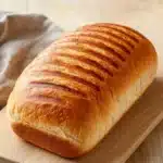 Grilled chicken sandwich bread - the image shows a freshly baked loaf of bread on a wooden cutting board. the bread is golden brown in color and has a soft, fluffy texture. it appears to be freshly baked, with visible ridges and grooves on the surface. the cutting board is placed on a light-colored wooden table, and there is a beige cloth napkin next to it. the background is blurred, but it seems to be a kitchen countertop.