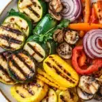 Grilled vegetables in air fryer - the image is a close-up of a plate of grilled vegetables. the plate is round and has a beige rim. the vegetables are arranged in a circular pattern on the plate. on the left side of the plate, there are sliced zucchini, yellow bell peppers, red onions, and mushrooms. the zucchinis are grilled to a golden brown color and have a crispy texture. the mushrooms are sliced into thin slices and are scattered throughout the plate with a sprig of parsley on top. the colors of the vegetables are vibrant and the plate is sitting on a white marble countertop.
