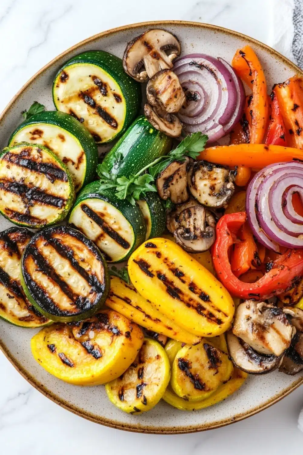 Grilled vegetables in air fryer - the image is a close-up of a plate of grilled vegetables. the plate is round and has a beige rim. the vegetables are arranged in a circular pattern on the plate. on the left side of the plate, there are sliced zucchini, yellow bell peppers, red onions, and mushrooms. the zucchinis are grilled to a golden brown color and have a crispy texture. the mushrooms are sliced into thin slices and are scattered throughout the plate with a sprig of parsley on top. the colors of the vegetables are vibrant and the plate is sitting on a white marble countertop.