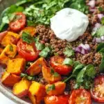 High protein beef and sweet potato bowl - the image is a close-up of a bowl of salad. the salad is made up of a bed of quinoa, diced tomatoes, sliced sweet potatoes, and red onions. the quinoa is a light brown color and appears to be seasoned with herbs and spices. the tomatoes are bright red and appear to be ripe and juicy. the sweet potatoes are cut into small cubes and are arranged in a pile on top of the quinoa. there are also some fresh greens, possibly arugula, scattered throughout the salad. on top, there is a dollop of sour cream. the bowl is sitting on a white marble countertop.