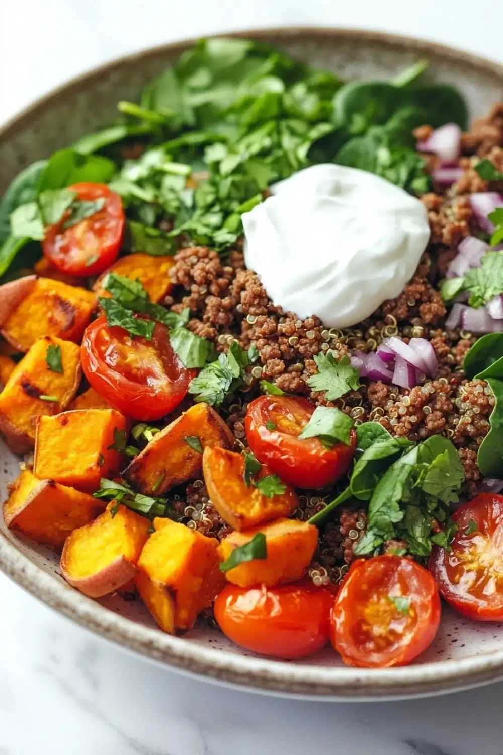 High protein beef and sweet potato bowl - the image is a close-up of a bowl of salad. the salad is made up of a bed of quinoa, diced tomatoes, sliced sweet potatoes, and red onions. the quinoa is a light brown color and appears to be seasoned with herbs and spices. the tomatoes are bright red and appear to be ripe and juicy. the sweet potatoes are cut into small cubes and are arranged in a pile on top of the quinoa. there are also some fresh greens, possibly arugula, scattered throughout the salad. on top, there is a dollop of sour cream. the bowl is sitting on a white marble countertop.