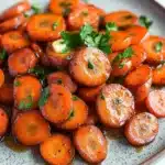 Honey and brown sugar glazed carrots - the image shows a plate of cooked carrots. the carrots are bright orange in color and appear to be seasoned with herbs and spices. they are arranged in a circular pattern on the plate, with some overlapping each other. the plate is white with a speckled pattern, and there are a few sprigs of parsley scattered around the plate. the background is blurred, but it appears to be a kitchen countertop.