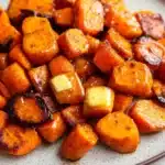 Honey glazed carrots and sweet potatoes - the image shows a plate of roasted sweet potatoes. the potatoes are orange in color and have a crispy texture. they are arranged in a circular pattern on the plate, with some overlapping each other. on top of the potatoes, there is a small block of butter, which is a light yellow color. the plate is white with a speckled pattern, and the background is blurred, making the potatoes the focal point of the image.