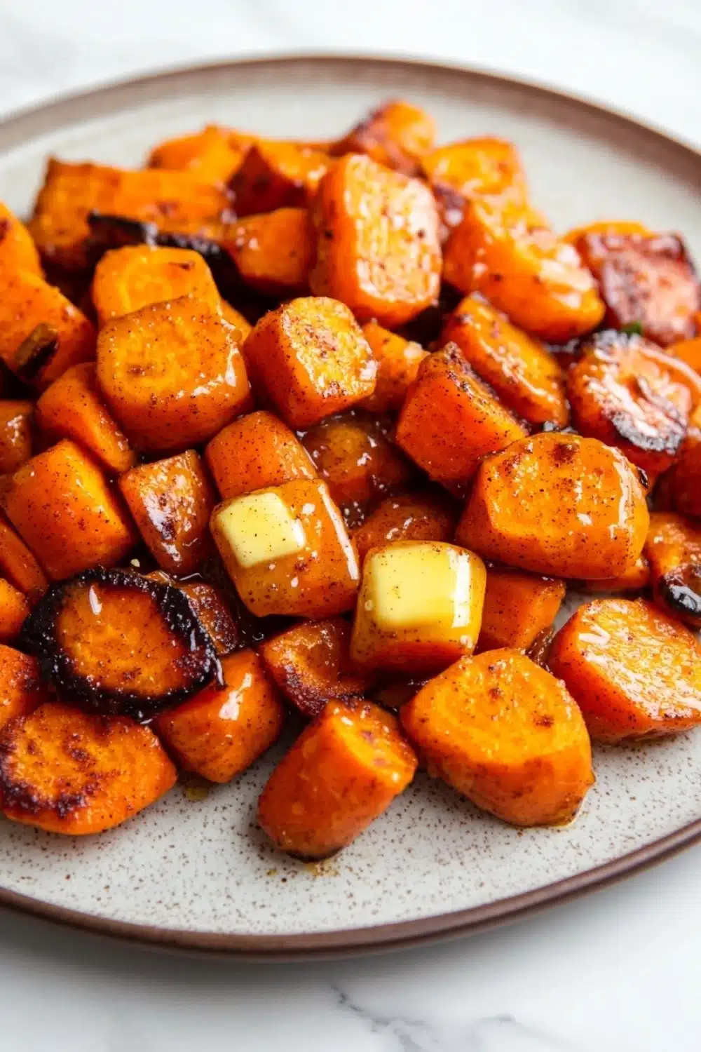 Honey glazed carrots and sweet potatoes - the image shows a plate of roasted sweet potatoes. the potatoes are orange in color and have a crispy texture. they are arranged in a circular pattern on the plate, with some overlapping each other. on top of the potatoes, there is a small block of butter, which is a light yellow color. the plate is white with a speckled pattern, and the background is blurred, making the potatoes the focal point of the image.