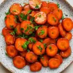 Honey glazed carrots vegan - the image is a close-up of a plate of cooked carrots. the carrots are bright orange in color and appear to be seasoned with herbs and spices. they are arranged in a circular pattern on the plate, with some overlapping each other. the plate is white with a speckled pattern around the edges. the background is a plain white surface. the overall appearance of the dish is fresh and appetizing.