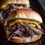 Hot roast beef sandwich slow cooker - the image shows three pulled pork sandwiches on a wooden tray. the sandwiches are made with a toasted bun and are topped with shredded beef, sliced onions, and melted cheese. the beef appears to be cooked medium-rare and the onions are caramelized. the cheese is melted and oozing out of the sides of the sandwiches. the tray is dark and the background is blurred, making the sandwiches the focal point of the image.