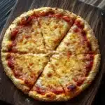 Keto pizza crust gluten free - the image shows a round pizza on a wooden cutting board. the pizza has a golden brown crust and is topped with melted cheese and red sauce. the cheese is melted and bubbly, and the sauce appears to be tomato-based. the cutting board is placed on a dark wooden table. the background is blurred, making the pizza the focal point of the image.