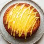 Lemon cake with lemon drizzle - the image shows a round cake on a gray plate with a white marble countertop. the cake appears to be a bundt cake with a golden brown crust and a drizzle of yellow icing on top. the icing is dripping down the sides of the cake, creating a smooth and glossy texture. the plate is round and has a speckled pattern around the edges. the background is blurred, making the cake the focal point of the image.