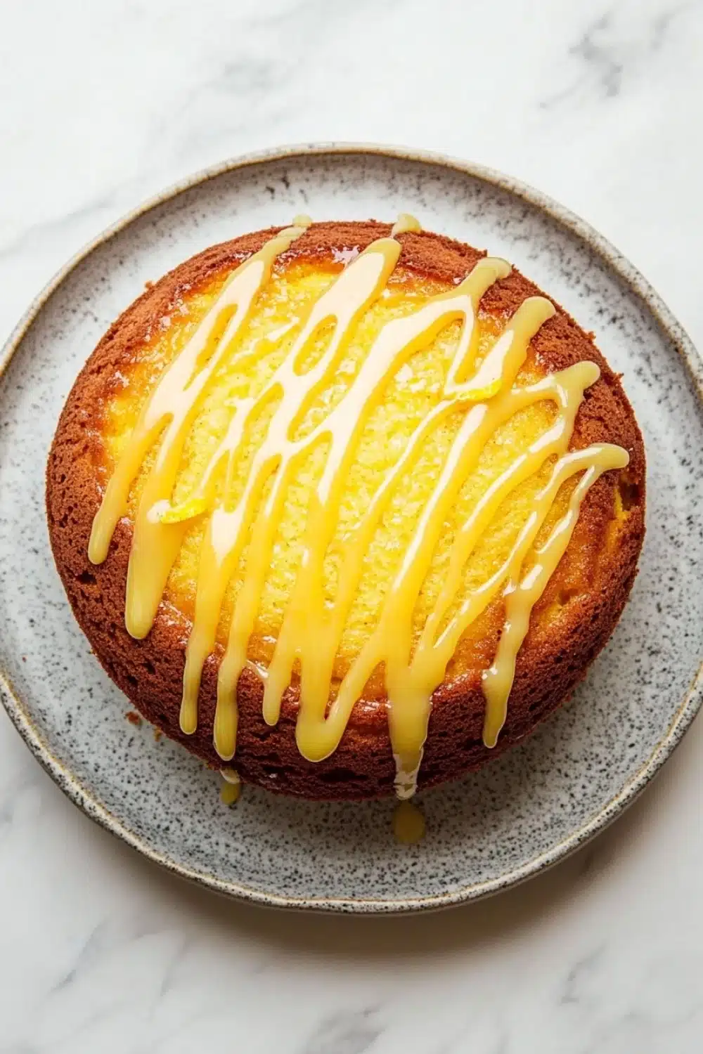 Lemon cake with lemon drizzle - the image shows a round cake on a gray plate with a white marble countertop. the cake appears to be a bundt cake with a golden brown crust and a drizzle of yellow icing on top. the icing is dripping down the sides of the cake, creating a smooth and glossy texture. the plate is round and has a speckled pattern around the edges. the background is blurred, making the cake the focal point of the image.