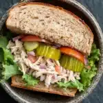 Loaded chicken salad sandwich - the image is a close-up of a sandwich in a bowl. the sandwich is made with two slices of toasted bread and is filled with a mixture of shredded chicken, diced tomatoes, pickles, and lettuce. the chicken appears to be seasoned with herbs and spices, and the vegetables are arranged in a colorful and appetizing manner. the bowl is made of dark brown ceramic with a speckled pattern around the edges. the background is a dark grey textured surface.