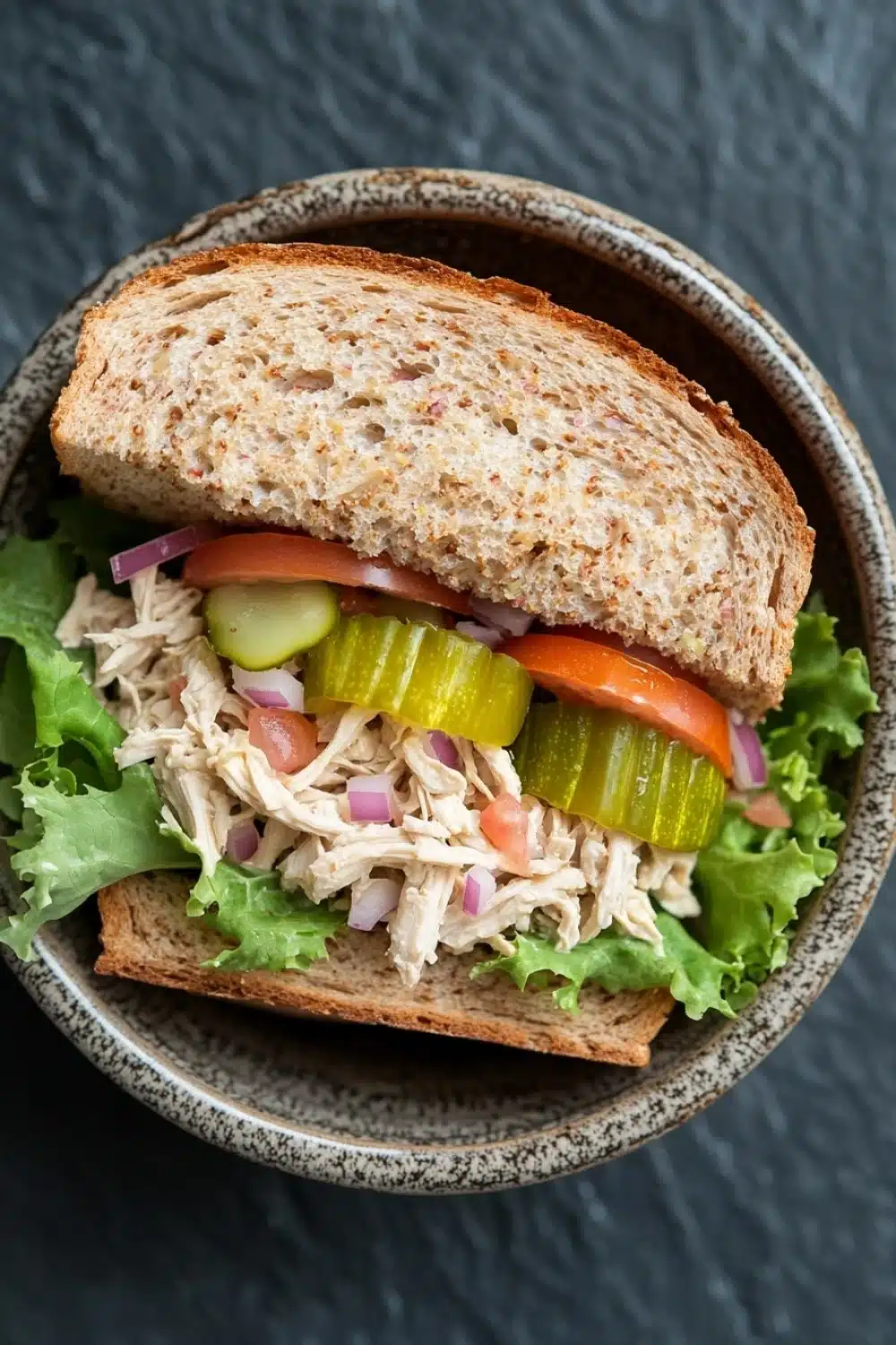 Loaded chicken salad sandwich - the image is a close-up of a sandwich in a bowl. the sandwich is made with two slices of toasted bread and is filled with a mixture of shredded chicken, diced tomatoes, pickles, and lettuce. the chicken appears to be seasoned with herbs and spices, and the vegetables are arranged in a colorful and appetizing manner. the bowl is made of dark brown ceramic with a speckled pattern around the edges. the background is a dark grey textured surface.