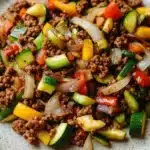 Low carb ground beef for dinner - the image is a close-up of a plate of food. the plate is round and has a light blue rim. the food appears to be a stir-fry with ground beef, sliced zucchini, red and yellow bell peppers, and sliced onions. the ground beef is dark brown and looks crispy on the outside. the vegetables are arranged in a colorful and appetizing manner, with some overlapping each other. the colors of the vegetables are vibrant and the dish looks fresh and ready to eat.