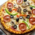 Low carb keto chicken crust pizza - the image is of a freshly baked pizza on a wooden cutting board. the pizza is round and has a golden brown crust. it is topped with melted cheese, sliced mushrooms, pepperoni, green bell peppers, and black olives. the toppings are arranged in a circular pattern on top of the cheese. the cutting board is placed on a white marble countertop with a gray and white striped napkin next to it.