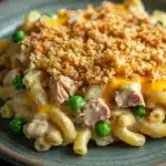 Lowfat tuna casserole - the image shows a plate of macaroni and cheese dish. the dish is served on a gray plate with a dark background. the top of the dish is covered in a crumbly, golden-brown breadcrumb topping, which appears to be a type of breadcrumbs. the cheese is melted and bubbly, and there are small chunks of ham and peas scattered throughout the dish. there are also a few green peas on the plate, adding a pop of color to the plate.