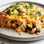 Make ahead chicken taco casserole - the image shows a slice of chicken enchilada casserole on a gray plate. the dish is made with shredded chicken, black beans, corn kernels, and crumbled cheese. the chicken is cooked to a golden brown color and appears to be seasoned with herbs and spices. the cheese is melted and bubbly, and there are small pieces of green onions scattered on top. the plate is garnished with a sprig of cilantro. the background is white, and the overall color scheme of the dish is warm and inviting.