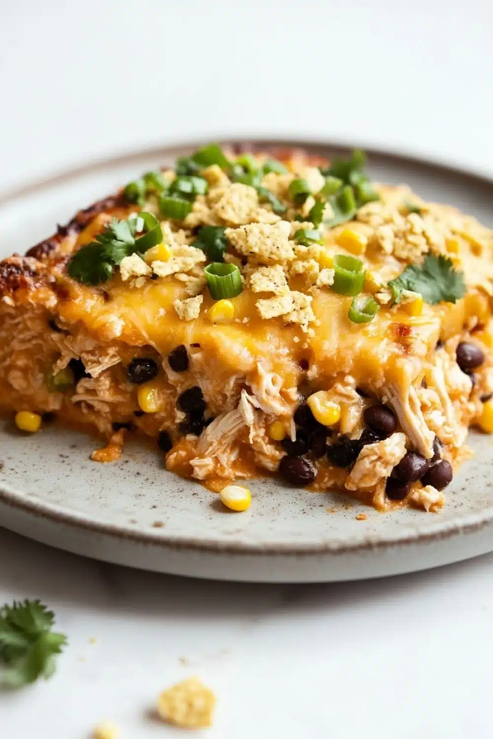 Make ahead chicken taco casserole - the image shows a slice of chicken enchilada casserole on a gray plate. the dish is made with shredded chicken, black beans, corn kernels, and crumbled cheese. the chicken is cooked to a golden brown color and appears to be seasoned with herbs and spices. the cheese is melted and bubbly, and there are small pieces of green onions scattered on top. the plate is garnished with a sprig of cilantro. the background is white, and the overall color scheme of the dish is warm and inviting.
