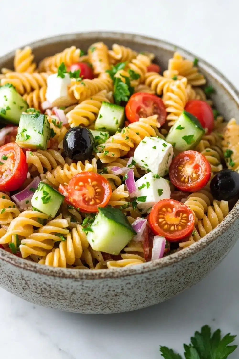 Make ahead italian pasta salad - the image is a close-up of a bowl of pasta salad. the bowl is made of ceramic and has a speckled pattern. the pasta is fusilli, which is a type of spiral-shaped pasta, and is mixed with various vegetables such as sliced tomatoes, cucumbers, red onions, and black olives. the vegetables are arranged in a colorful and appetizing manner, with some of them overlapping each other. there are also some fresh cilantro leaves scattered throughout the salad, adding a pop of green color to the dish. the background is a white marble countertop.