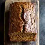Moist pumpkin zucchini bread - the image shows a loaf of bread on a wooden cutting board. the bread appears to be freshly baked and has a golden brown crust on top. it has a crumbly texture and is filled with zucchini seeds. the cutting board is placed on a dark textured surface, possibly a table or countertop. the background is blurred, making the bread the focal point of the image.