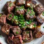 My incredible juicy garlic butter steak bites - the image is a close-up of a plate of food. the plate is made of ceramic and has a light brown color with a speckled texture. on top of the plate, there are several pieces of cooked steak, which appear to be medium-rare and have a dark brown color. the steak is garnished with chopped parsley and a drizzle of white sauce. the sauce is drizzled over the steak, giving it a glossy sheen. the dish looks appetizing and ready to eat.